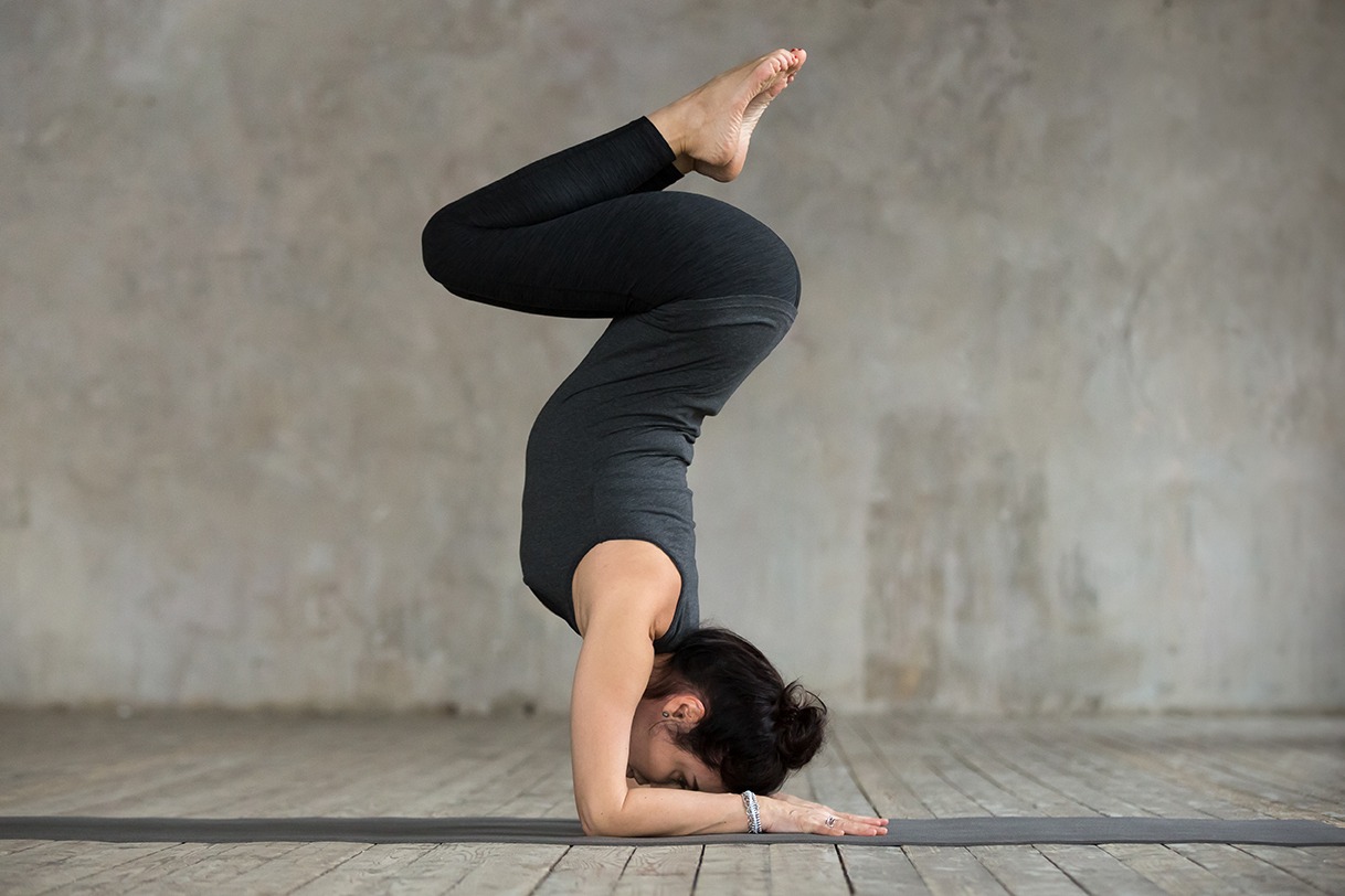 Young sporty woman practicing yoga, doing Pincha Mayurasana exercise, handstand pose, working out, wearing sportswear, black pants and top, indoor full length, gray wall in yoga studio
